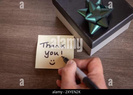 Eine Person schreibt eine Dankesnotiz auf ein Blatt Papier. Die Notiz ist mit schwarzem Marker und neben einer Geschenkbox mit einem Band geschrieben. Stockfoto