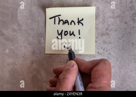 Eine Person schreibt eine Dankesnotiz auf ein Blatt Papier. Die Notiz befindet sich auf einem Holztisch und die Person schreibt sie mit einem Marker. Szene ist positiv Stockfoto