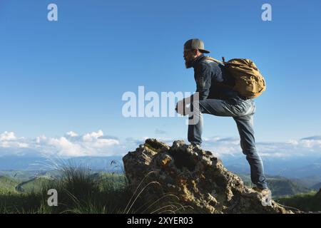 Stilvoller bärtiger Reisender in Sonnenbrille und einer Mütze mit Rucksack in Denim-Anzug und gelben Schuhen steht auf einem großen Stein vor dem Hintergrund Stockfoto