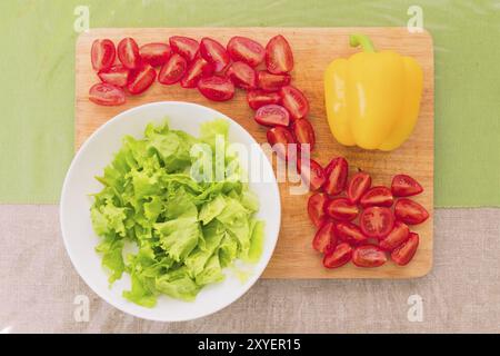 Frisch gehackte grüne Salatblätter liegen auf einem Holzschneidebrett neben der gelben Paprika und gehackten roten Kirschtomaten auf einem grünen Tisch. Die Stockfoto