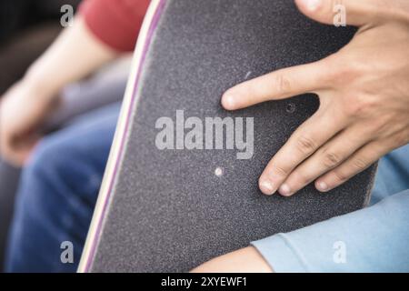 Ein kleiner Junge auf den Knien klebt das Griptape in Begleitung von Freunden bei sonnigem Wetter auf ein Skateboard. Vorbereitung eines Skateboards für einen Skatepark-Wettkampf Stockfoto