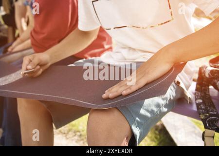 Ein kleiner Junge auf den Knien klebt das Griptape in Begleitung von Freunden bei sonnigem Wetter auf ein Skateboard. Vorbereitung eines Skateboards für einen Skatepark-Wettkampf Stockfoto