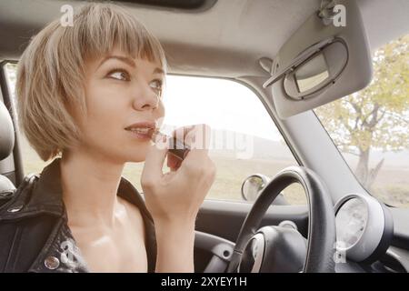 Niedliche blonde Frau mit kurzen Haaren, die Lippen färben, während sie am Steuer ihres Autos sitzt. Das Konzept, eine Frau zu fahren. Unangemessene Fahrstunden. D Stockfoto