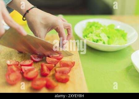 Die Hände eines jungen Mädchens hacken die Kirschtomaten auf einem hölzernen Schneidebrett auf einem grünen Tisch in einem Wohnbereich gegen einen Teller mit geschnittenen Salatblättern Stockfoto