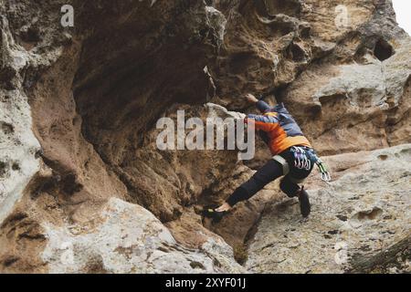 Hipster, Kletterer im Alter, in dem man einen wunderschönen Felsen ohne Versicherung und Helm hochklettert. Ein Bergsteiger in Hut und Daunenjacke mit Tasche für magnes Stockfoto