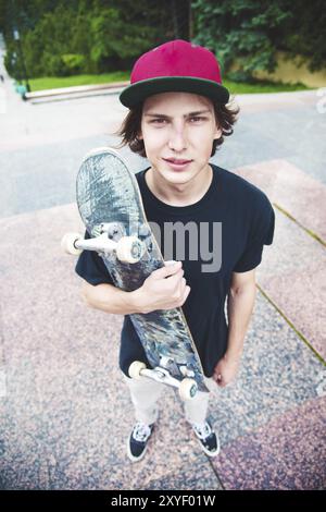 Ein junger langhaariger Skater in T-Shirt und durchgehender Cap steht mit einem Skateboard vor dem Hintergrund der Stadt Stockfoto