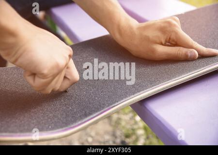 Ein kleiner Junge auf den Knien klebt das Griptape in Begleitung von Freunden bei sonnigem Wetter auf ein Skateboard. Vorbereitung eines Skateboards für einen Skatepark-Wettkampf Stockfoto