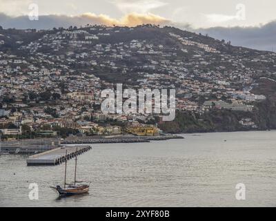 Blick auf eine Küstenstadt mit Hügelhäusern und Boot im Vordergrund in der Abenddämmerung, madeira, portugal Stockfoto