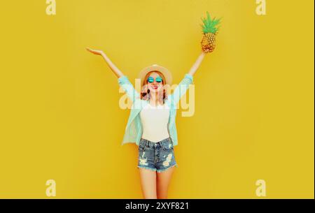 Glückliche fröhliche Frau hebt ihre Hände mit Ananas hoch, viel Spaß im Sommerhut auf gelbem Hintergrund Stockfoto