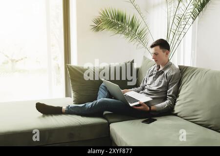 Junger Mann sitzt auf dem grünen Sofa mit grauem Laptop, schwarzem Telefon. Der Mann hält ein silbernes Notizbuch, liest, tippt etwas. Arbeit von zu Hause, Bildung Co Stockfoto