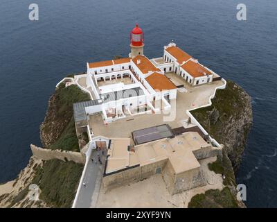 Ein roter Leuchtturm auf einem Felsen über dem Meer mit mehreren weißen Gebäuden, die ein ruhiges Gefühl vermitteln, aus der Vogelperspektive, Leuchtturm, Cabo de Sao Vicente, Kap St. V. Stockfoto