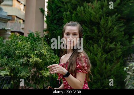 Eine rothaarige Frau in einem roten Blumenkleid tanzt fröhlich mit ihrem Handy in der Hand und hält das Wesen eines sonnigen Tages fest, während sie in die Kamera blickt. Stockfoto