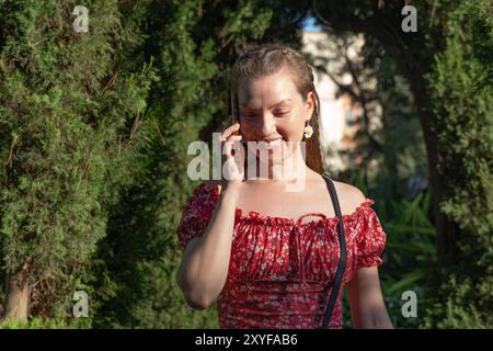 Eine fröhliche junge rothaarige Frau mit geflochtenen Haaren, die ein rotes Blumenkleid trägt, lächelt, während sie in einem üppig grünen Park mit ihrem Smartphone plaudert. Stockfoto