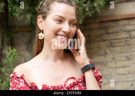 Eine junge rothaarige Frau in einem roten Sommerkleid wird an einem sonnigen Tag auf ihrem Smartphone bei einem angenehmen Gespräch beobachtet. Stockfoto