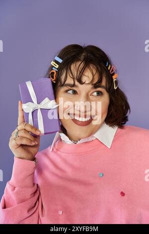 Eine Frau mit bunten Haarspangen lächelt aufgeregt, während sie eine lila Geschenkbox hält. Stockfoto