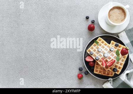 Stapel von zuckerpulverförmigen Waffeln mit Honig auf einem Teller auf dem Handtuch und grauen Beton- oder Schiefertisch mit Heidelbeere, gehackten Erdbeeren und Minzblättern Stockfoto