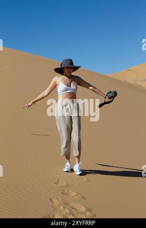 Eine weiße Frau, die auf den Wüstendünen läuft, mit Hut und Kamera in der Hand. Stockfoto