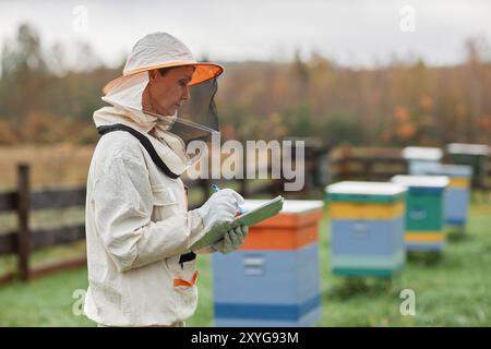 Seitenansicht einer Reifen Imkerin im Bienenschutzanzug, die in Notizblock schreibt, während sie um bunte Holzstöcke auf der Herbstbienenfarm arbeitet, Kopierraum Stockfoto