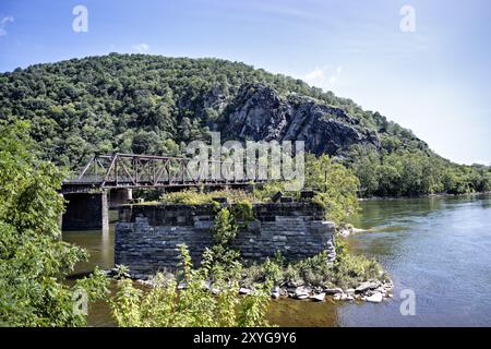 HARPERS FERRY, West Virginia – Blick von der Lower Town Harpers Ferry über den Potomac River in Richtung Maryland Heights, mit der historischen Eisenbahnbrücke und den Überresten der Bollman Bridge Pylone im Vordergrund. Diese Szene fängt die strategische Bedeutung dieser historischen Flussüberquerung und ihre Rolle in der amerikanischen Transportgeschichte ein. Stockfoto