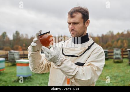 Taille oben Schuss eines ernsten erwachsenen männlichen Imkers im Bienenschutzanzug, der ein Glas mit rohem Honig gegen den grauen Herbsthimmel auf der Bienenfarm hält, Kopierraum Stockfoto