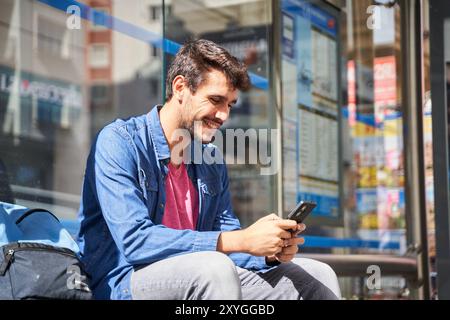 Bärtiger junger Mann in Denim-Hemd, der an der Bushaltestelle sitzt und auf sein Handy schaut, während er im Hintergrund der modernen Stadt auf den Bus wartet Stockfoto