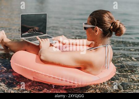 Eine Frau sitzt auf einem pinkfarbenen Schlauchboot im Wasser und benutzt einen Laptop. Das Konzept der Entspannung und Freizeit, während die Frau ihre Zeit am genießt Stockfoto
