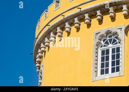 Pena-Palast Gelbe Fassade Verziertes Fenster Maurische Details Sintra Portugal | SINTRA, Portugal — Die leuchtend gelbe Fassade des Pena-Palastes rahmt ein verziertes Fenster mit maurischen Details, ein Detail, das die Philosophie des Palastes der bewussten stilistischen Juxtaposition einfängt. Dieser Abschnitt gehört zum Flügel des Neuen Palastes, der gelb gestrichen ist, um sich vom tiefen Rot des älteren Klosterblocks abzuheben — ein kühnes polychromatisches Schema, das Ferdinand II entwarf und Parques de Sintra später wiederherstellte, nachdem die Farben im Laufe des 20. Jahrhunderts verblasst waren. Die hier sichtbaren neo-maurischen und neo-manuelinischen Elemente wurden zwischen Stockfoto