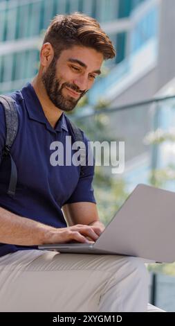 Lächelnder bärtiger, männlicher Freiberufler, der am Laptop arbeitet und in der Nähe von Wolkenkratzern sitzt Stockfoto