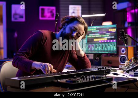 Müder Künstler verliert den Schlaf, um neue Klaviernoten im Heimstudio zu lernen, spielt elektronische Tastatur des Midi-Controllers, um Songs zu komponieren. Der Produzent ist erschöpft und stresst, während er an Musik arbeitet. Stockfoto