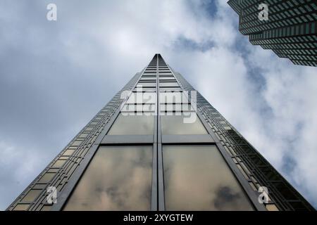 Ein hoch aufragender Wolkenkratzer erstreckt sich zu einem bewölkten Himmel, dessen Glasfassade die Atmosphäre widerspiegelt. Die Perspektive betont ihre beeindruckende Höhe. Stockfoto