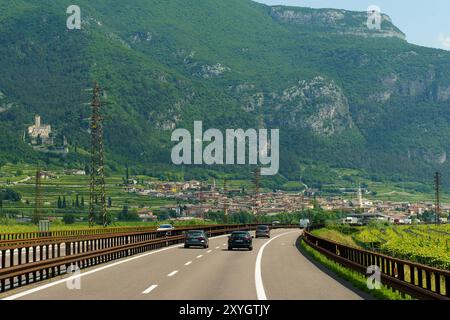 Avio, Italien - 8. Juni 2023: Autos fahren auf einer Autobahn, die sich durch ein Tal schlängelt, mit einer kleinen Stadt in der Ferne und einem majestätischen Berg Stockfoto