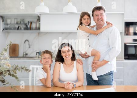 Mom sitzt in der Küche neben dem Sohn, Dad steht hinter ihr mit einer Vorschultochter in Armen Stockfoto