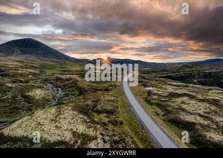 Aus der Vogelperspektive auf eine Straße, die sich durch die sanften Hügel schlängelt, den Sonnenuntergang hinter den Bergen, der einen warmen Glanz über die beschauliche nordische Landschaft wirft Stockfoto