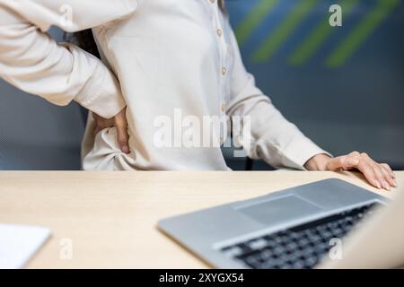 Büroangestellter fühlt sich unwohl, während er am Schreibtisch sitzt und Rückenschmerzen hat. Die Person drückt auf den unteren Rücken, um die Belastung oder Muskelschmerzen zu lindern. Das Bild zeigt häufige arbeitsbezogene Gesundheitsprobleme. Stockfoto
