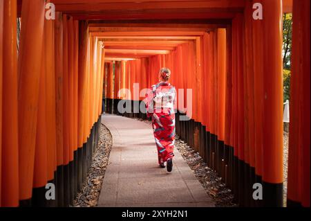 Eine Frau in einem traditionellen roten Kimono spaziert durch einen Tunnel aus orangefarbenen Torii-Toren an einem japanischen Schrein. Die Szene fängt die heitere Schönheit von ein Stockfoto