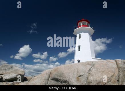 Ein unberührter weißer Leuchtturm mit einer leuchtend roten Spitze steht hoch auf felsigem Gelände vor einem tiefblauen Himmel mit flauschigen weißen Wolken. Stockfoto