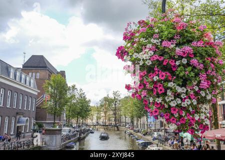 Großer hängender Blumentopf mit bunten Blumen vor einem Kanal und einer Brücke unter leicht bewölktem Himmel, Amsterdam, Niederlande Stockfoto