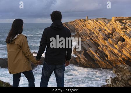 Ein Paar kaukasische Frauen und Männer, die Hände halten und auf Baleal Island, Peniche, Portugal, Europa, die Klippenlandschaft mit Meereswellen betrachten Stockfoto