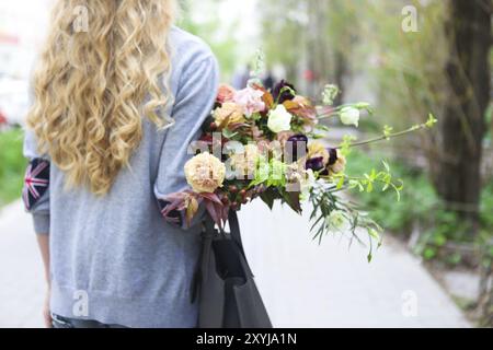 Junge, schöne Frau, die auf der Straße läuft mit Tasche und Blumenstrauß. Model mit stilvoller Kleidung und Accessoires Stockfoto