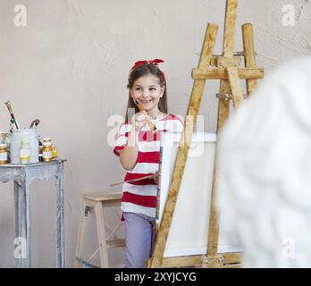 Kleine Künstler Mädchen hält einen Pinsel mit Blick auf eine Leinwand auf einer Staffelei. Konzept der Kreativität und Spaß Stockfoto