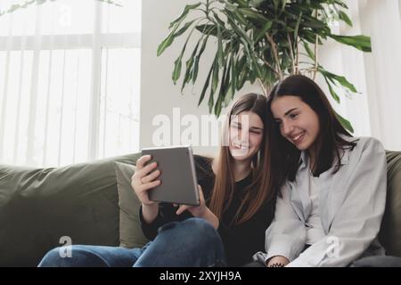 Zwei Teenager-Schwesternmädchen sitzen auf dem grünen Sofa im Wohnzimmer und sehen sich ein lustiges Video am Tablet an, lächelnd und lachend. Schöne Leute, frend Stockfoto