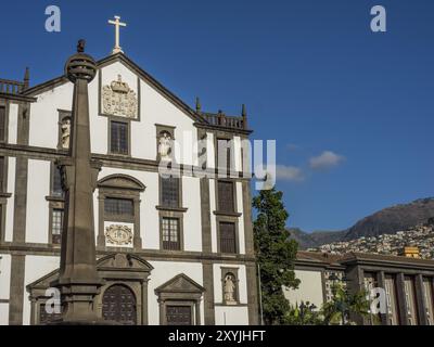 Kirche mit Kreuz und Denkmal im Vordergrund unter blauem Himmel und Berge im Hintergrund, madeira, portugal Stockfoto