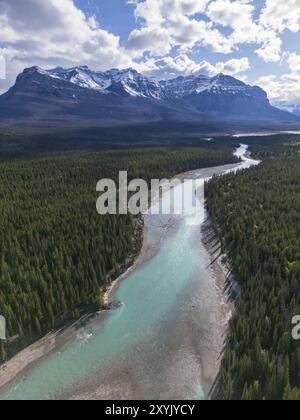 Ein türkisfarbener Fluss, der sich durch einen dichten grünen Wald schlängelt, inmitten schneebedeckter Berge unter einem klaren Himmel mit Wolken, in den kanadischen Rockies, Alberta Stockfoto