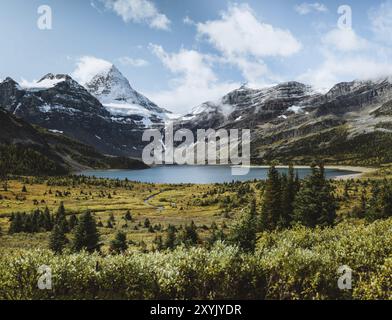 Eine unberührte Berglandschaft mit einem klaren See, umgeben von schneebedeckten Gipfeln und üppiger grüner Vegetation unter einem hellblauen Himmel mit verstreutem Clou Stockfoto