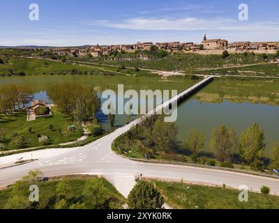 Ein Dorf entlang eines Flusses mit Brücke und Straße, umgeben von grüner Landschaft und Bäumen unter blauem Himmel, aus der Vogelperspektive, Maderuelo, Rio Riaza, Fluss R Stockfoto