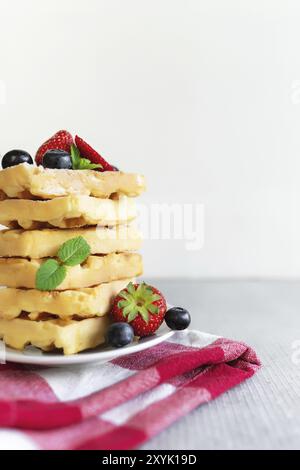 Weißer Teller auf rotem Küchentuch mit hausgemachten belgischen Waffeln, Heidelbeere, Erdbeerschnitt, Minzblatt, Zuckerpulver auf grauem Hintergrund Stockfoto