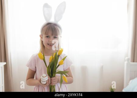 Osterkonzept. Kleines lächelndes Mädchen in weißen flauschigen Hasenohren und rosa Kleid mit gelben Tulpenblüten und bunten Eiern. Zimmereinrichtung. Stockfoto