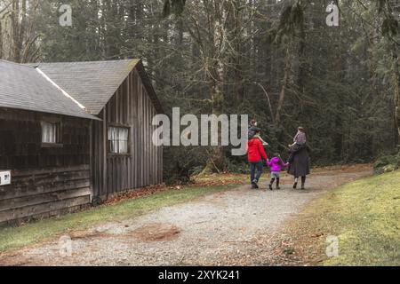 Eine Familie spaziert zusammen auf einem Schotterweg vorbei an einer rustikalen Holzhütte in einem Wald, warm gekleidet für die Wintersaison Vancouver Island, BC, Kanada, Nein Stockfoto