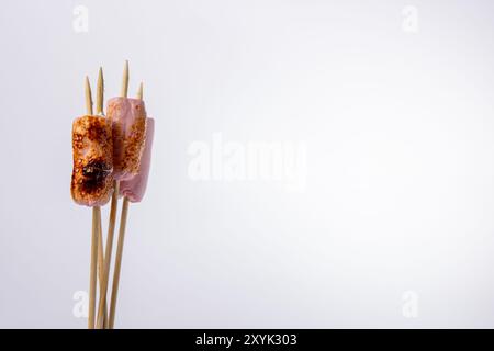 Nahaufnahme von Spießen mit gerösteten Marshmallows auf der linken Seite vor einem strahlend weißen Hintergrund. Das Foto hebt die goldbraune Textur des hervor Stockfoto