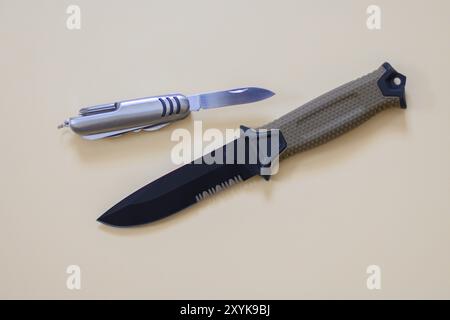 Ein Jagdmesser mit beigefarbenem Griff liegt neben einem Schweizer Armeemesser auf cremefarbener Oberfläche Stockfoto